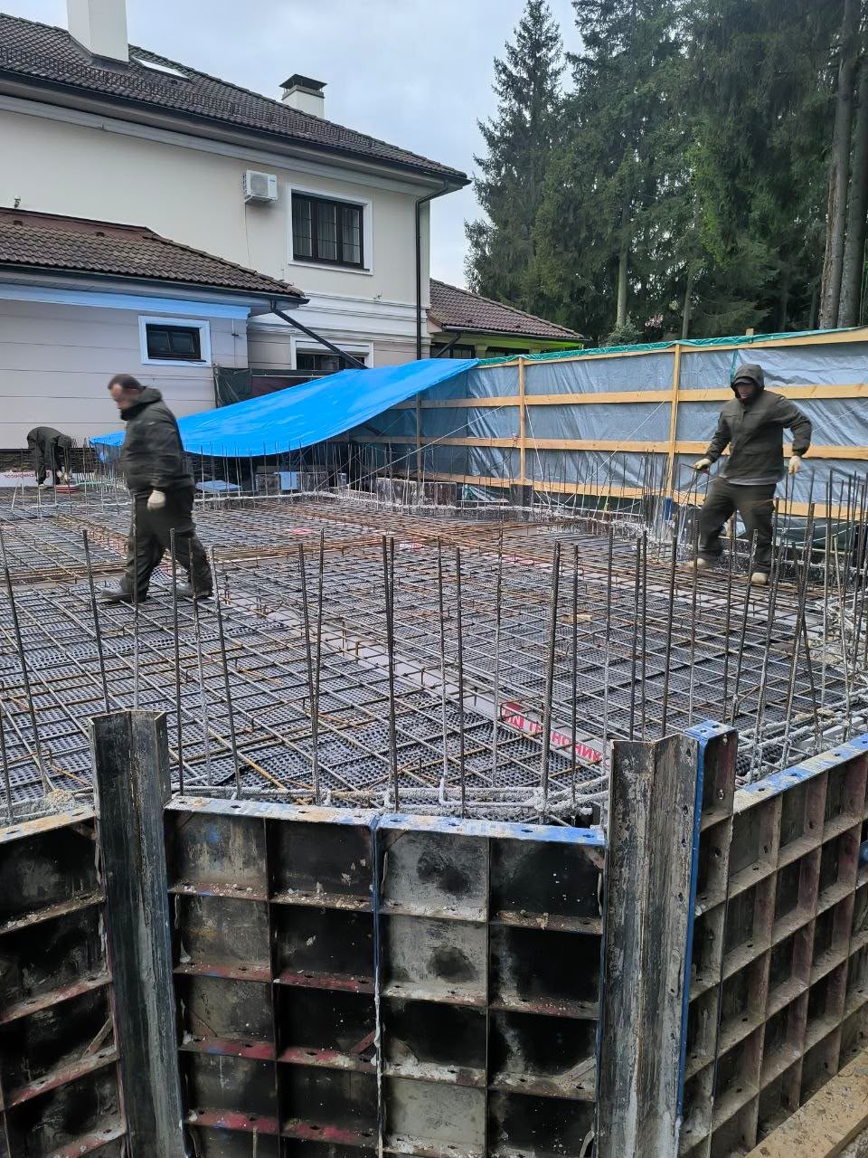 Арматурный каркас плиты в металлической опалубке. На каркасе двое рабочих. За забором уже стоящий дом и ели.